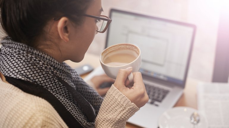 Eine Frau sitzt am Laptop und hält eine Tasse Kaffee in der Hand.