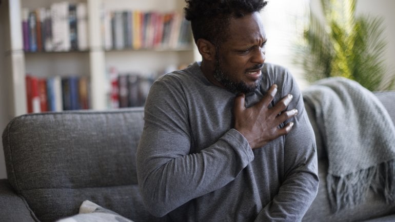 Mann mit Herzrhythmusstörungen sitzt auf Sofa und fasst sich an die Brust.