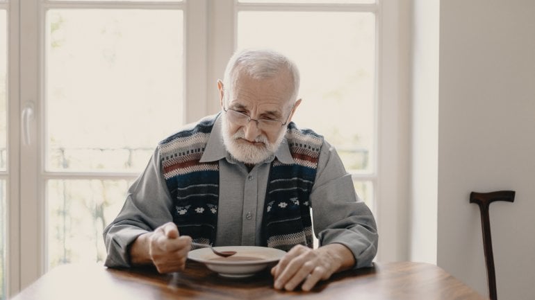 Älterer Mann mit Mangelernährung sitzt allein am Tisch und isst Suppe.
