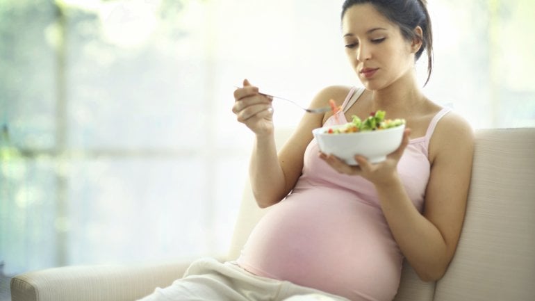 Eine schwangere Frau sitzt auf einem Sofa und isst Salat.
