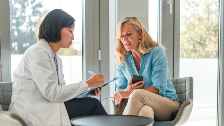 Eine Ärztin im weißen Kittel sitzt gemeinsam mit einer Frau mittleren Alters und spricht mit ihr.