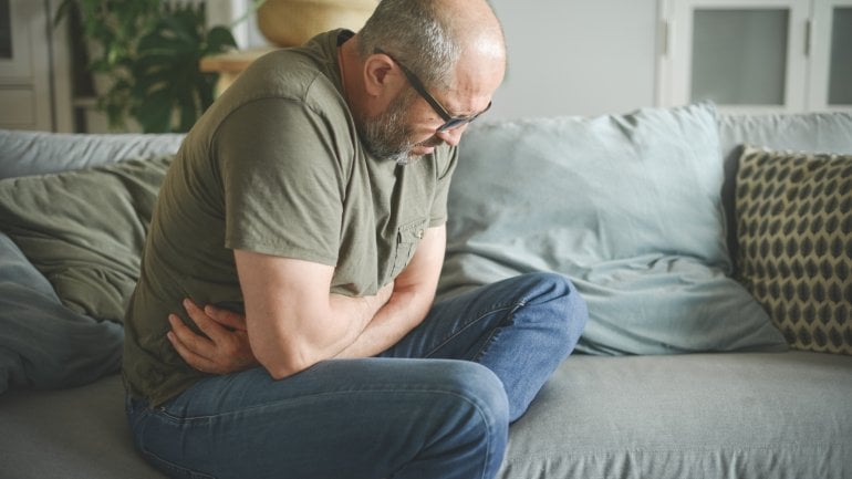 Mann mit Kurzdarmsyndrom sitzt auf Sofa und hält sich vor Schmerzen den Bauch.