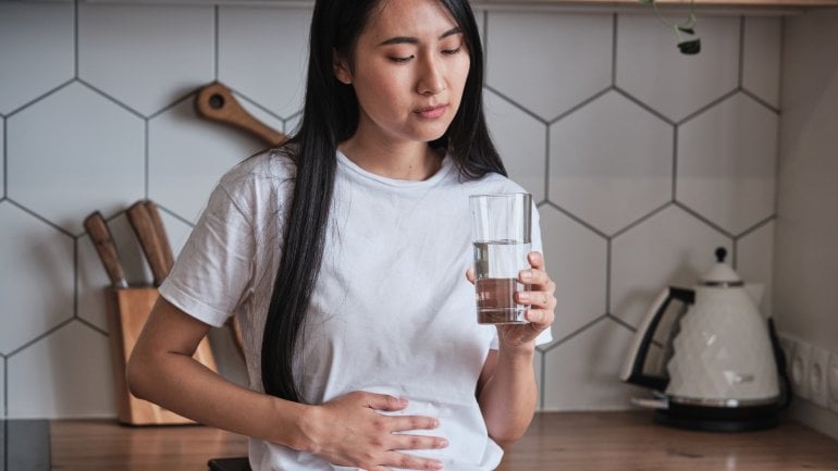 Junge, schwarzhaarige Frau mit Magenschmerzen steht in der Küche mit einem Glas Wasser in der Hand