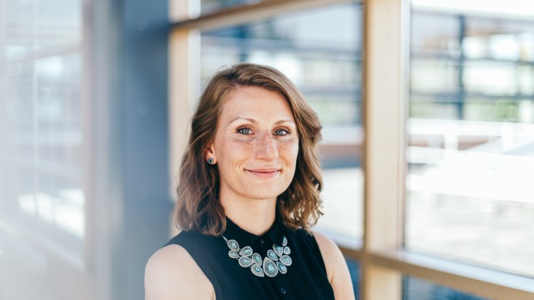 Frau mit hellbraunen Locken steht im Büroflur und lächelt in die Kamera