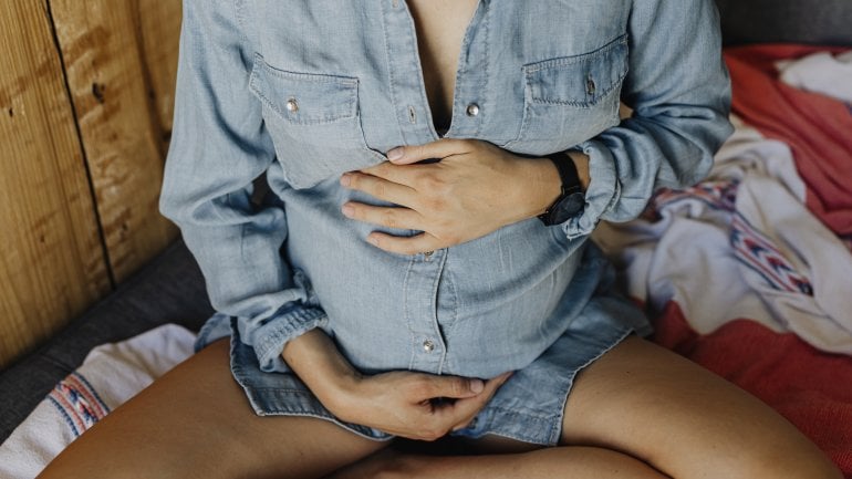 Man sieht nur den schwangeren Bauch einer Frau mit Jeanskleid und ihre Hände darauf