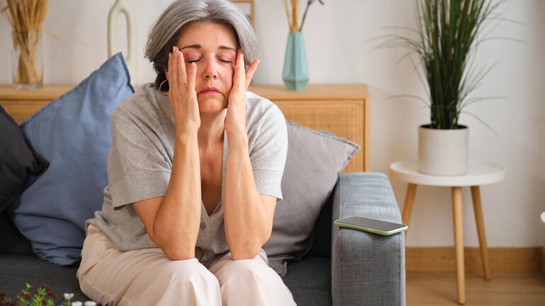 Frau sitzt müde auf dem Sofa und fasst sich mit Händen an den Kopf.