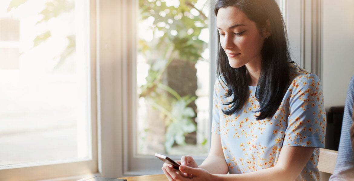 Junge, dunkelhaarige Frau sitzt am Tisch und schaut lächelnd auf ihr Smartphone, das sie in den Händen hält