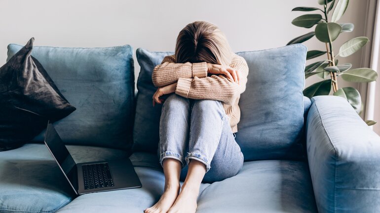 Eine junge Frau sitzt auf dem Sofa. Sie wirkt erschöpft und belastet und hat ihren Kopf auf ihre verschränkten Arme gelegt.