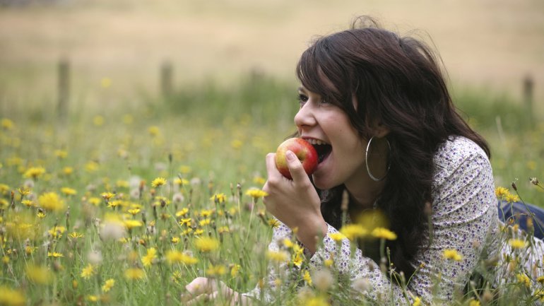 Eine junge Frau liegt in einer Blumenwiese und beißt in einen Apfel.