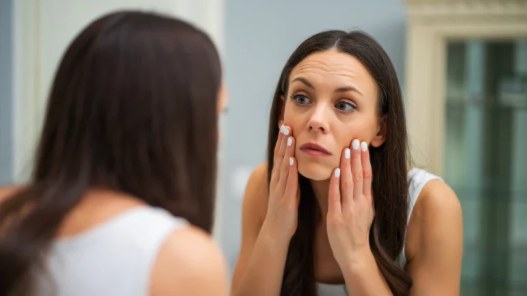 Eine Frau betrachtet sich im Spiegel und berührt ihr Gesicht mit den Händen.