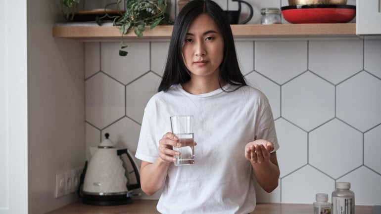 Eine junge Frau steht in der Küche, in der einen Hand ein Glas Wasser, in der anderen eine Tablette