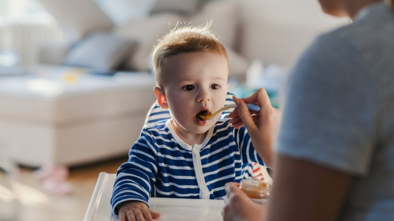 Das Bild zeigt einen kleinen Jungen, der von seiner Mutter mit Babybrei gefüttert wird.