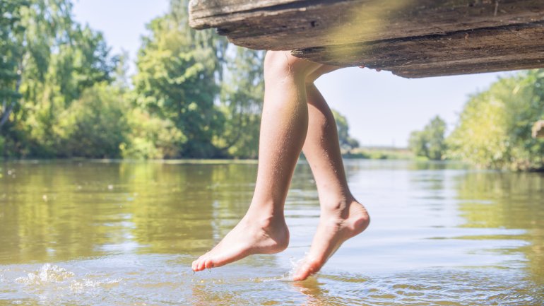 Jemand sitzt auf einem hölzernen Steg an einem See und hält seine Zehen ins Wasser.