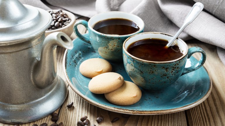Neben einer silbernen Kaffeekanne steht ein Teller mit zwei Tassen Kaffee und ein paar Plätzchen.