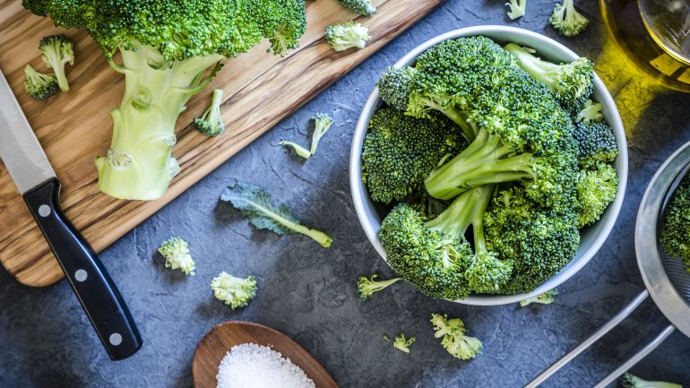 Brokkoli-Röschen in einer Schale, ein Brokkoli-Kopf auf einem Holzbrett un dein Holzlöffel mit Salz auf einem blauen Tisch