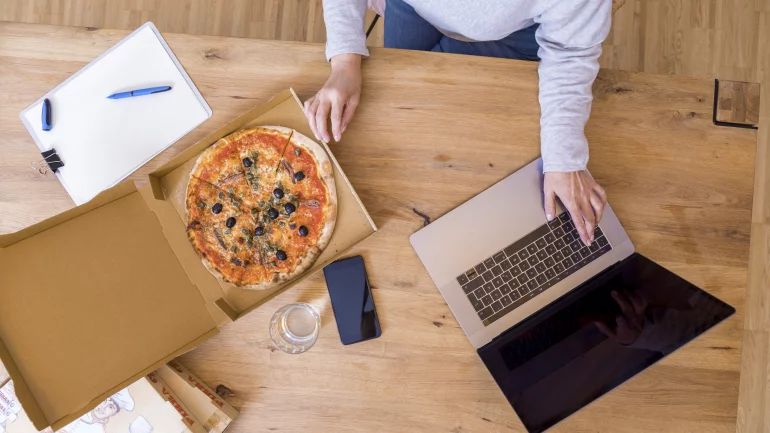 Eine Person sitzt mit Laptop und Pizza am Schreibtisch. 