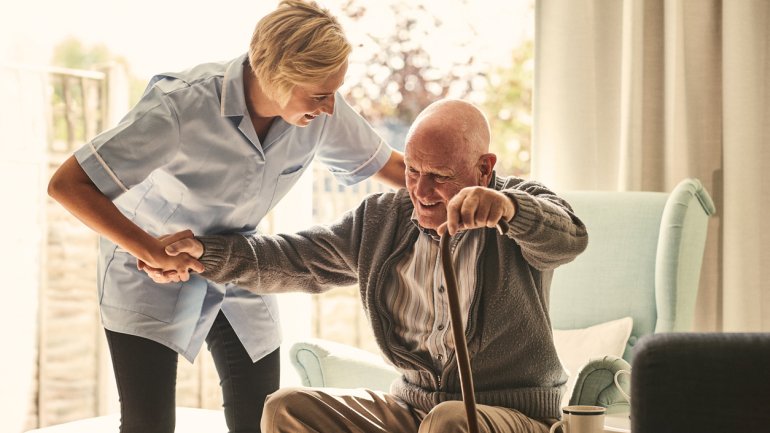 Eine Pflegerin hilft einem älteren Mann dabei, aus dem Sessel aufzustehen.