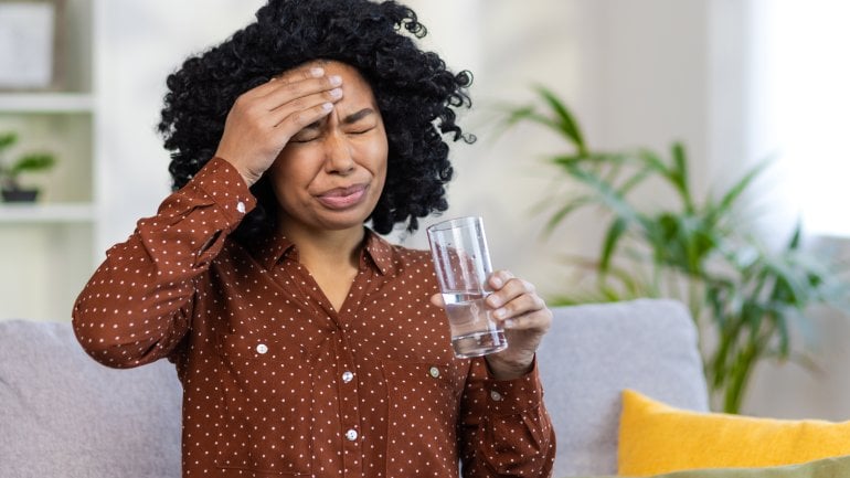 Eine Frau trinkt ein Glas Wasser und hält sich den schmerzenden Kopf
