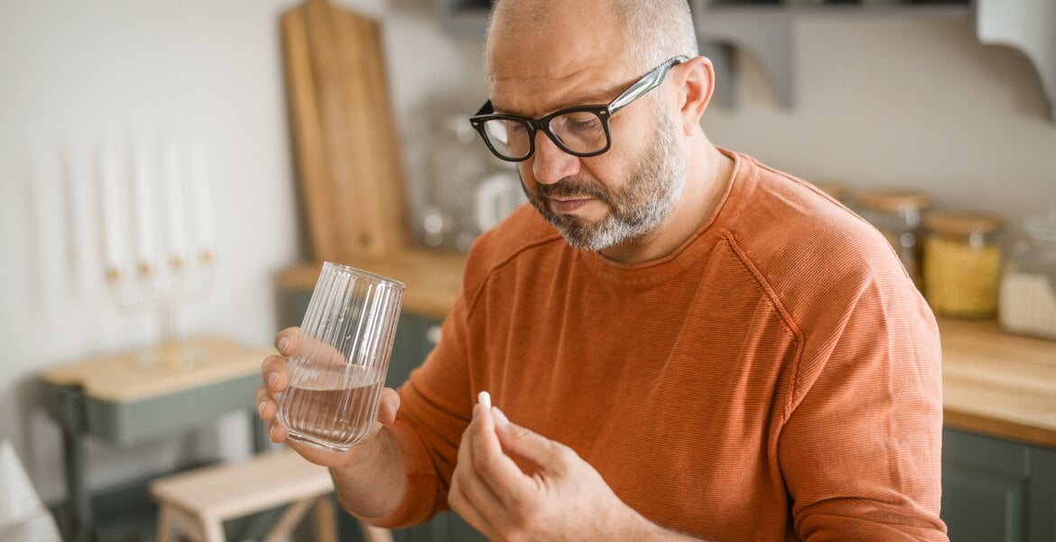Mann mittleren Alters in orangefarbigem Pullover nimmt eine Tablette ein, in der anderen Hand hält er ein Wasserglas