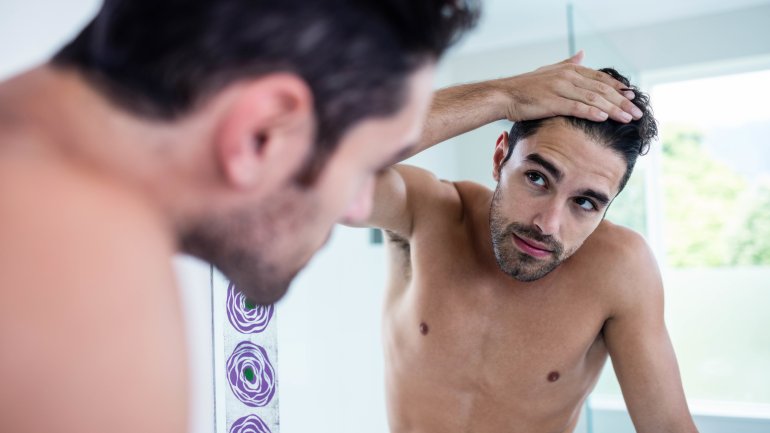 Das Bild zeigt einen jungen Mann, der vor einem Spiegel steht und seinen Haaransatz betrachtet.