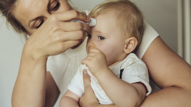 Drei-Tage-Fieber: Mutter hält einem Baby ein Fieberthermometer an die Stirn
