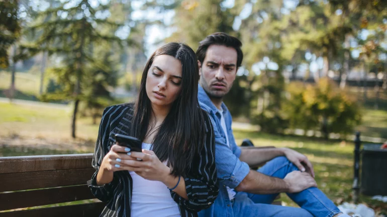 Ein Paar sitzt auf einer Bank. Der Mann schaut eifersüchtig auf das Handy seiner Freundin.