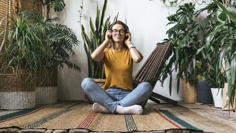 Eine Frau sitzt mit Kopfhörern zwischen Pflanzen auf dem Boden und entspannt sich.