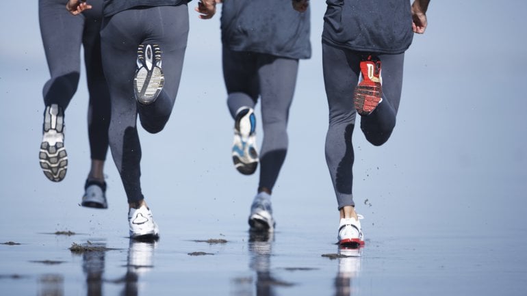 Man sieht die Bein von vier Läufern, die am Strand joggen, von hinten.