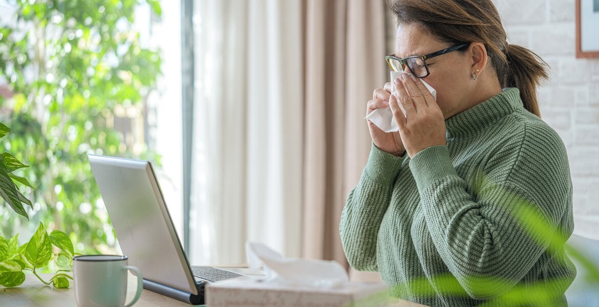 Frauin grünem Pullover arbeitet im Büro am Laptop und putzt sich die Nase mit einem Taschentuch