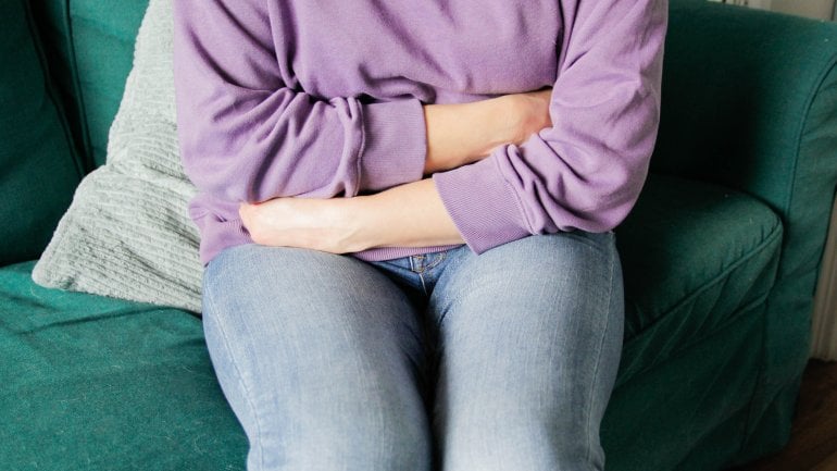Frau mit Gebärmuttersenkung sitzt auf Sofa und hält sich Arme vor den Unterleib.