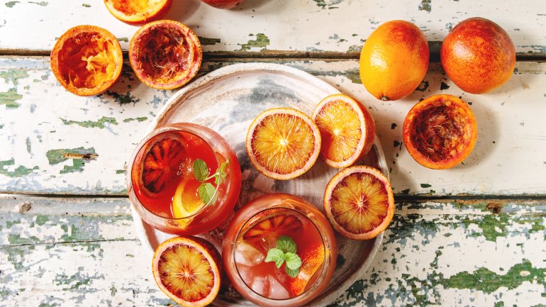 Man sieht zwei Cocktails auf einem weißen Tisch, daneben liegen Grapefruits und Passionsfrüchte.