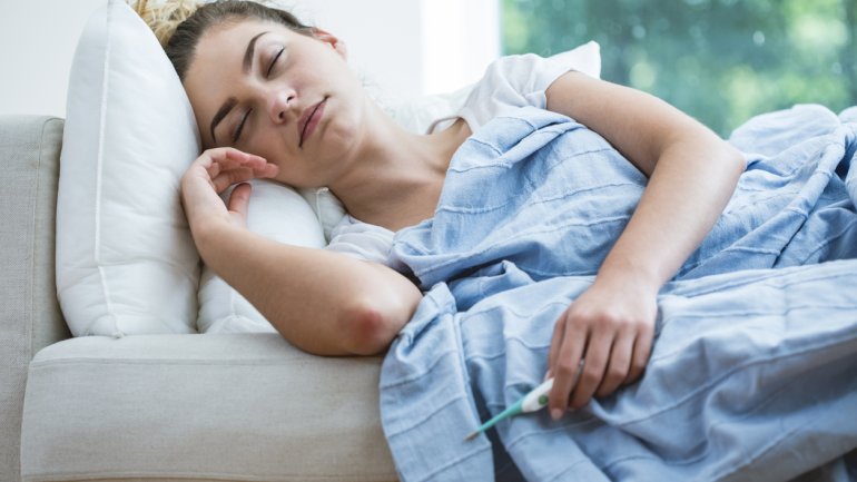 Eine krank und erschöpft wirkende Frau liegt zugedeckt auf einem Sofa und hält ein Fieberthermometer in der Hand.