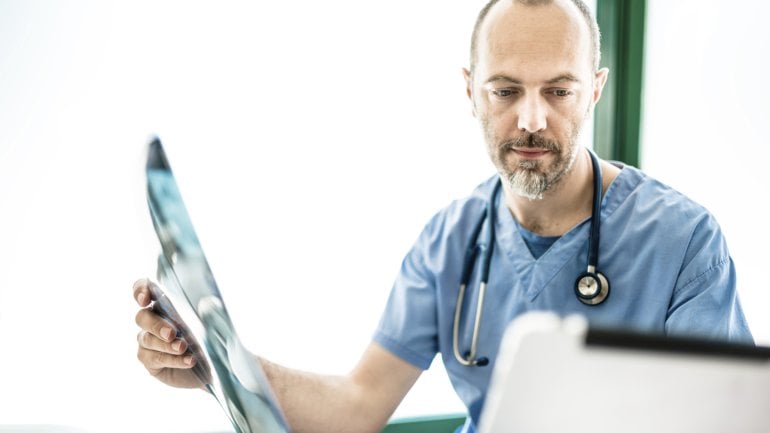 Ein Arzt hält ein Rötgenbild in der rechten Hand und blickt nachdenklich auf ein Notepad.
