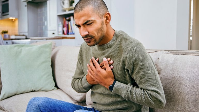 Junger Mann mit Herzmuskelentzündung sitzt auf Couch und hält sich Hand auf schmerzende Brust.