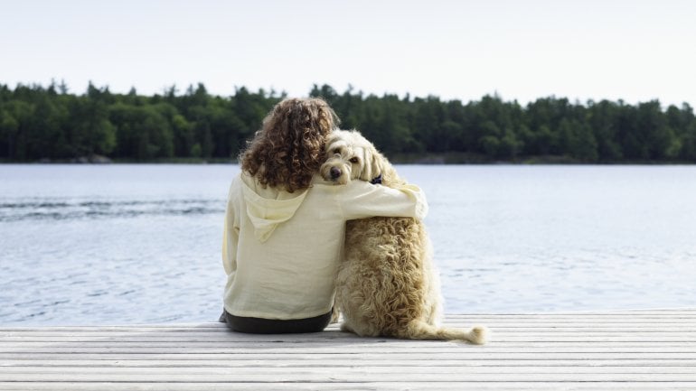 Eine Frau und ein Hund sitzen nebeneinander auf einem Holzsteg an einem Gewässer; die Frau hat den Arm um den Hund gelegt.