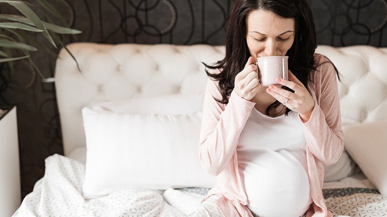 Eine Schwangere sitzt im Bett und trinkt Tee