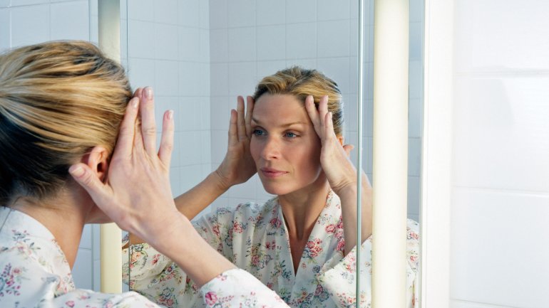 Eine Frau betrachtet sich im Spiegel und strafft ihr Gesicht mit den Händen.