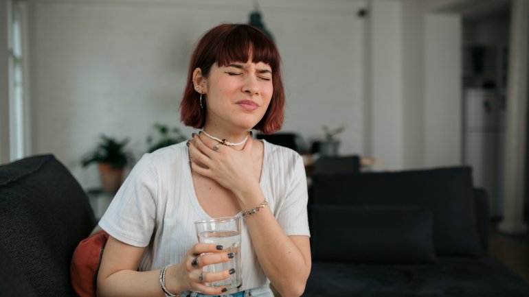Eine Frau sitzt mit einem Glas Wasser auf der Couch und fasst sich an den kratzenden Hals