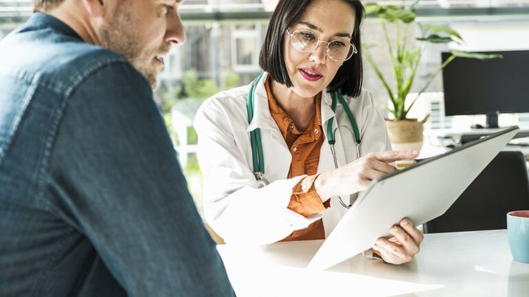 Braunhaarige Ärztin mit Brille erklärt ihrem Patienten den Befund auf einem Tablet in der Hand