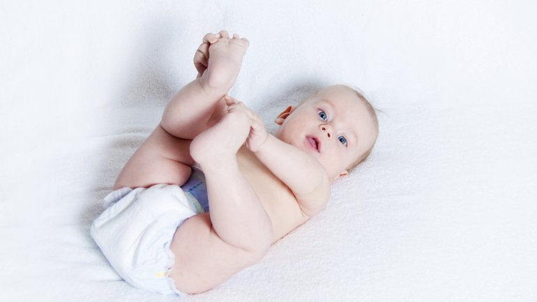 Etwa fünf Monate altes Baby liegt in Windeln auf dem Rücken und spielt mit seinen Füßen.