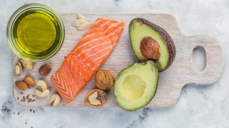 Bild von Fisch, Öl, Nüssen und Avocado auf einem Tisch, die viel Omega-3-Fettsäuren enthalten.