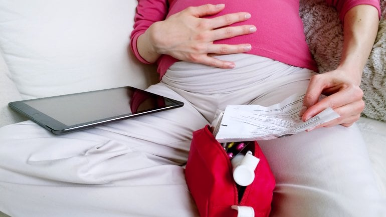 Eine Schwangere sitzt mit Tablet und Medizintasche auf dem Sofa und liest einen Beipackzettel.