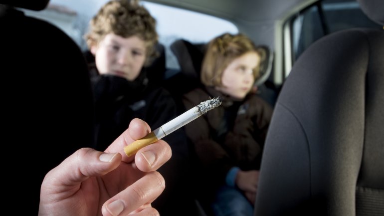 Zwei auf dem Rücksitz eines Autos sitzende Kinder sind dem Zigarettenqualm des rauchenden Beifahrers ausgesetzt.