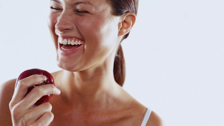 Man sieht eine lachende junge Frau mit einem Apfel in der Hand. 