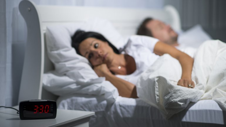 Eine Frau liegt neben einem schlafenden Mann wach im Bett; ihr Wecker zeigt 3:30 Uhr an.