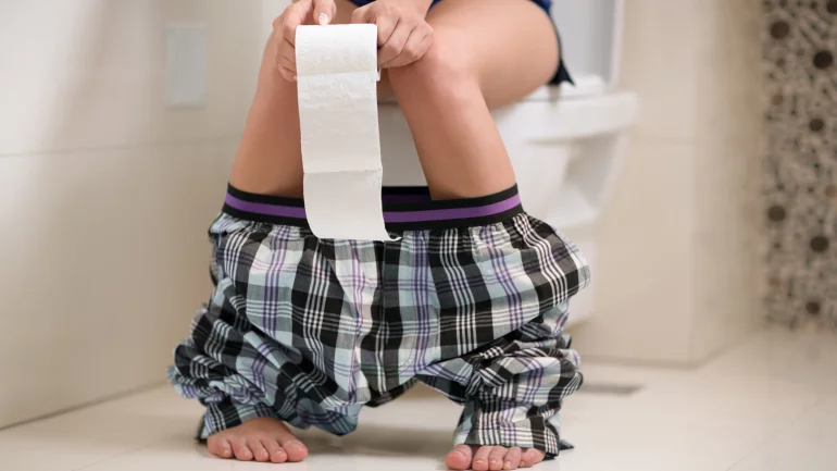 Eine Frau sitzt auf der Kloschüssel und hält eine Rolle Toilettenpapier in den Händen.