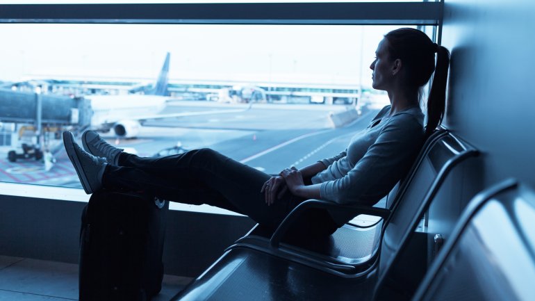 Eine Frau sitzt im Flughafenterminal mit Blick auf das Vorfeld auf einer Bank und hat ihre Füße auf einem Koffer abgelengt.