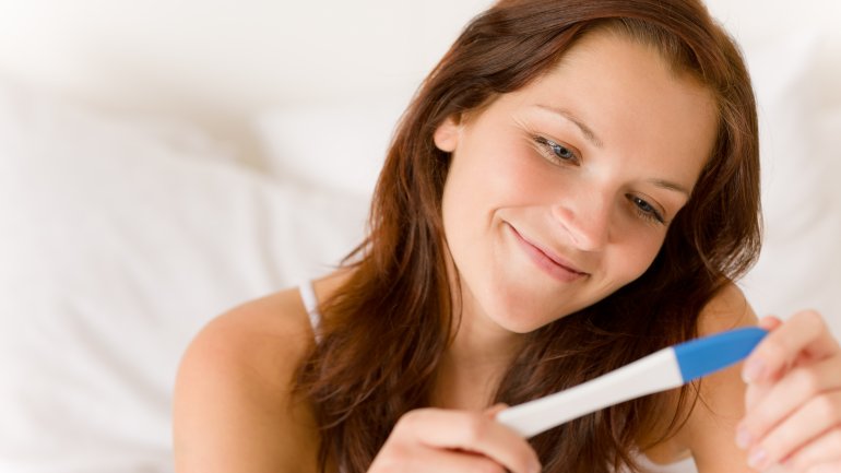 Das Bild zeigt eine lächelnde Frau, die auf einen Schwangerschaftstest schaut.