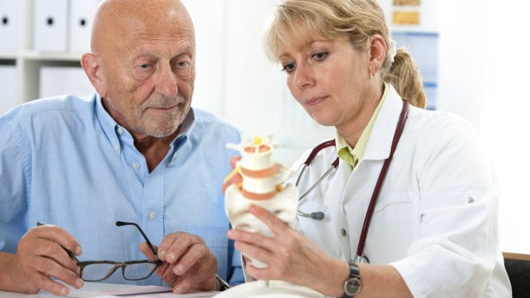 Ein älterer Mann sitzt neben einer Ärztin, die einen Teil eines Wiebelsäulenmodells in der Hand hält und mit der anderen Hand darauf deutet.