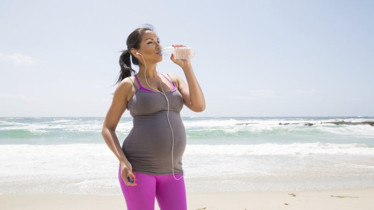 Das Bild zeigt eine Schwangere am Strand mit einer Flasche Wasser in der Hand.